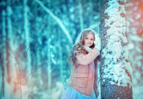 Beautiful little girl with long hair. Little girl in the winter forest. girl in a white knitted scarf. Girl in a Zara sheepskin coat. The little girl smiles. Snow. A little girl is standing by a tree.