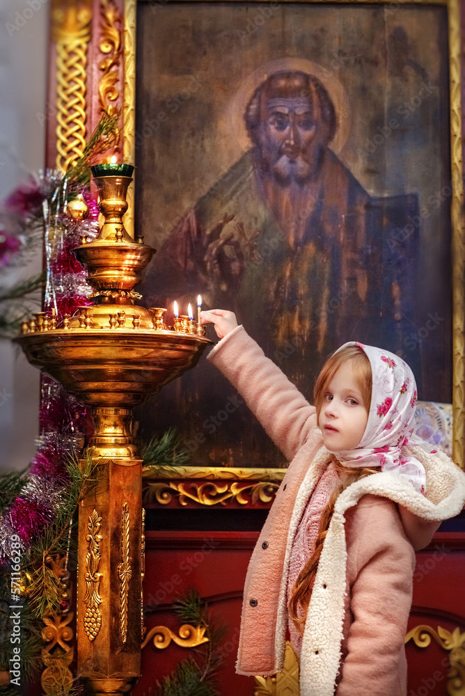 Fototapeta premium A little beautiful girl puts candles in the temple in the church. Orthodox holiday. Christian holiday. Prayer. Icons