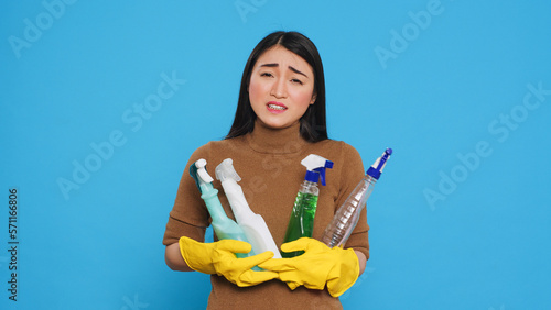 Fotografija Overwhelmed tired housekeeper wearing gloves while holding multiple sanitary cleanliness products while cleaning house for clients