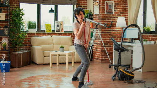 Cheerful girlfriend listening to music on headset and mopping floors, sweep dirt. Housewife feeling happy and cleaning household with tools and appliances, enjoying dance moves.