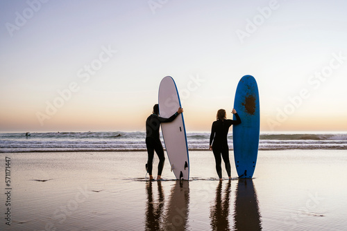 Couple of anonymous surfers on the shore of the beach at sunset standing with their boards looking at the horizon while studying the incoming wave series.