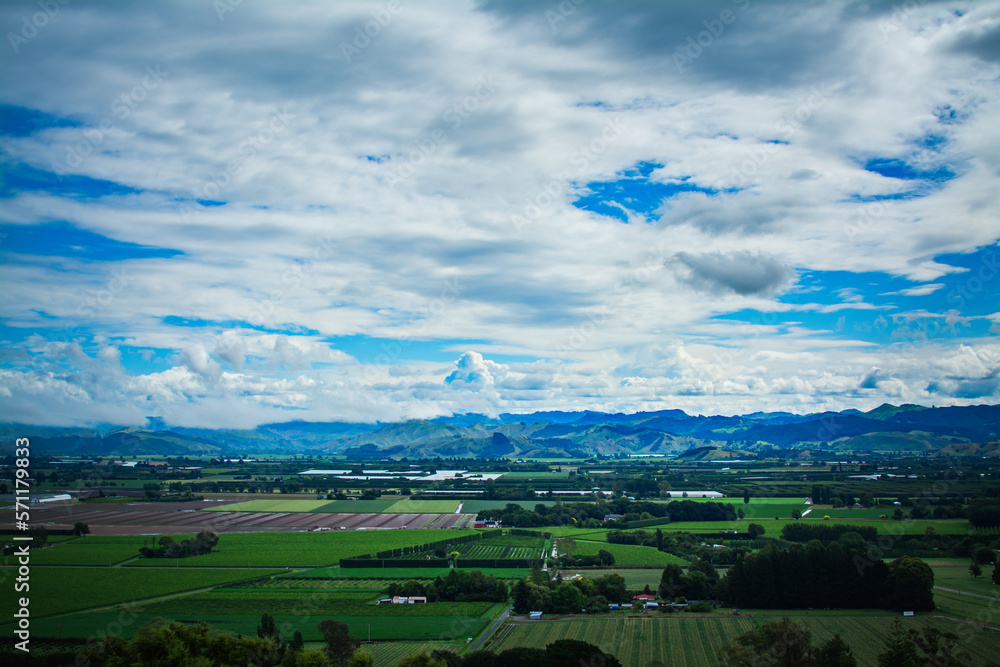Aerial view over lush green farmland, distand mountain range rising over horizon under cloudy sky. Breathtaking New Zealand Landscape. Greys Hill Lookout, Gisborne, North Island, New Zealand