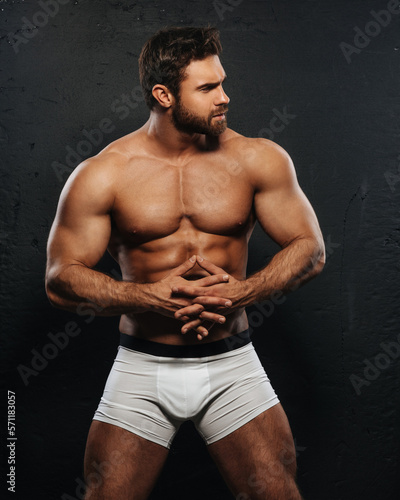 Naked muscled man in white boxer underwear on black background. Sexy bearded guy in studio. Muscular male model holding his hands together in front of him.