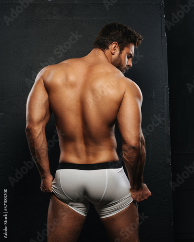 Naked muscled man in white boxer underwear on black background. Sexy back of bodybuilder in studio. Backside of muscular male model holding his underwear.