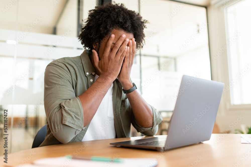 Tired young african-american male employee suffering from severe headache migraine. Weary male freelancer man overwhelmed with computer work, closes his face with both hands Tired young african-american male employee suffering from severe headache migraine. Weary male freelancer man overwhelmed with computer work, closes his face with both hands