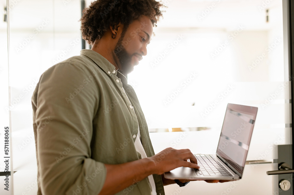 Ambitious highly-skilled handsome curly african-american male employee using laptop in modern office, businessman stands in hallway and typing, side view Ambitious highly-skilled handsome curly african-american male employee using laptop in modern office, businessman stands in hallway and typing, side view