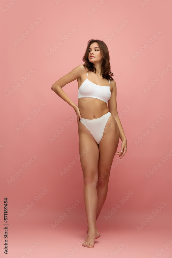 Young tall thin girl with long soft brown hair wearing white inner wear posing over pink color background. Concept of natural beauty, body and skin care, healthy eating Young tall thin girl with long soft brown hair wearing white inner wear posing over pink color background. Concept of natural beauty, body and skin care, healthy eating