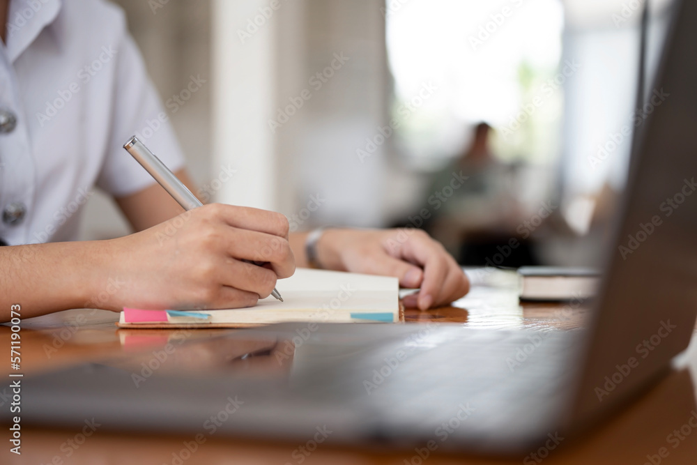 Young female student writing on notebook, online learning via laptop computer sitting in library