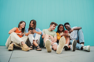 Multiracial group of young friends enjoying and smiling using their mobile phone app sitting at teal wall. Diverse teenagers having fun sharing messages with each other on cellphone at blue clor