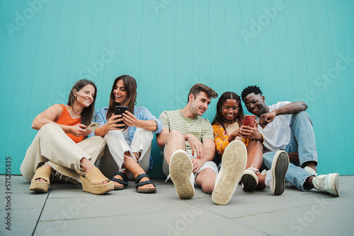 Fotografiet Multiracial group of young friends enjoying and smiling using their mobile phone app sitting at teal wall