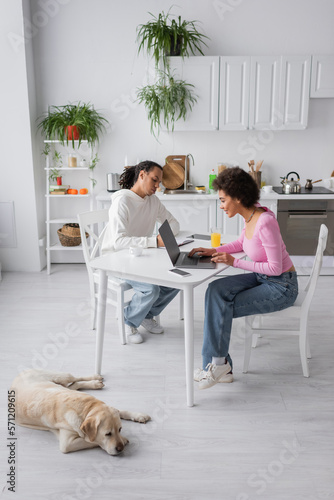 Smiling african american woman using laptop near boyfriend and labrador in kitchen.