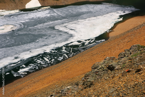 Frozen highland lake among dramatic mountain red and orange rocks with snow Altai Siberia Russia