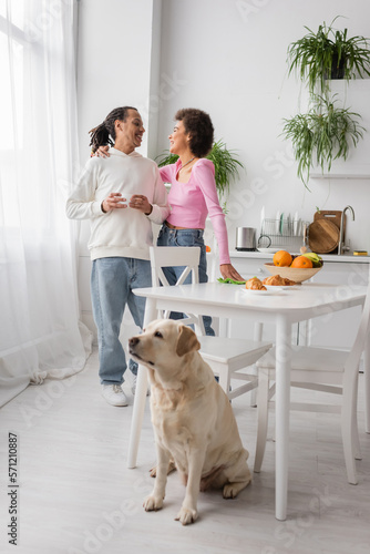 African american woman hugging boyfriend with coffee near labrador in kitchen.