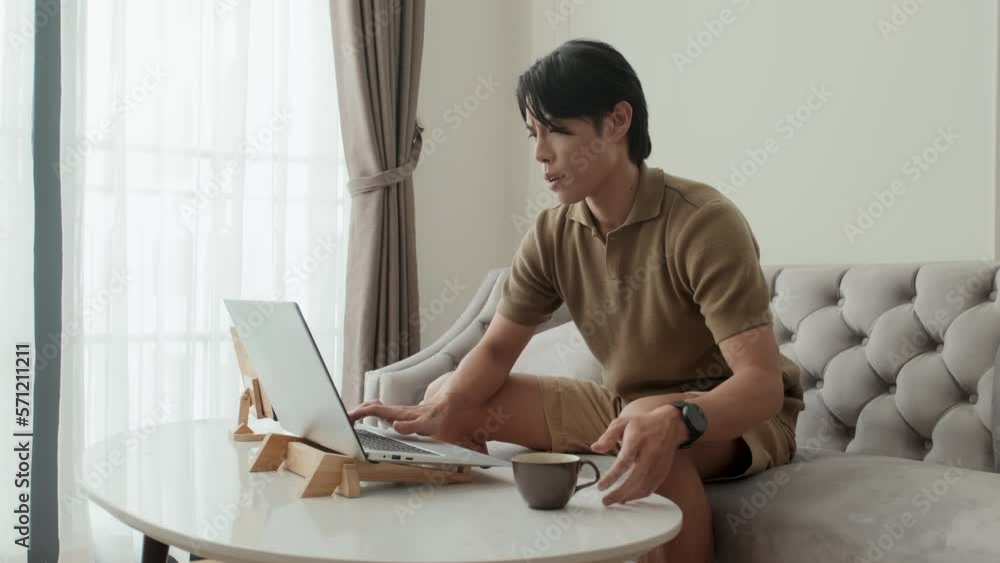 Young Asian businessman or male freelancer in casualwear typing on laptop keyboard and having tea while sitting on grey soft comfortable couch by white oval table in living room Young Asian businessman or male freelancer in casualwear typing on laptop keyboard and having tea while sitting on grey soft comfortable couch by white oval table in living room