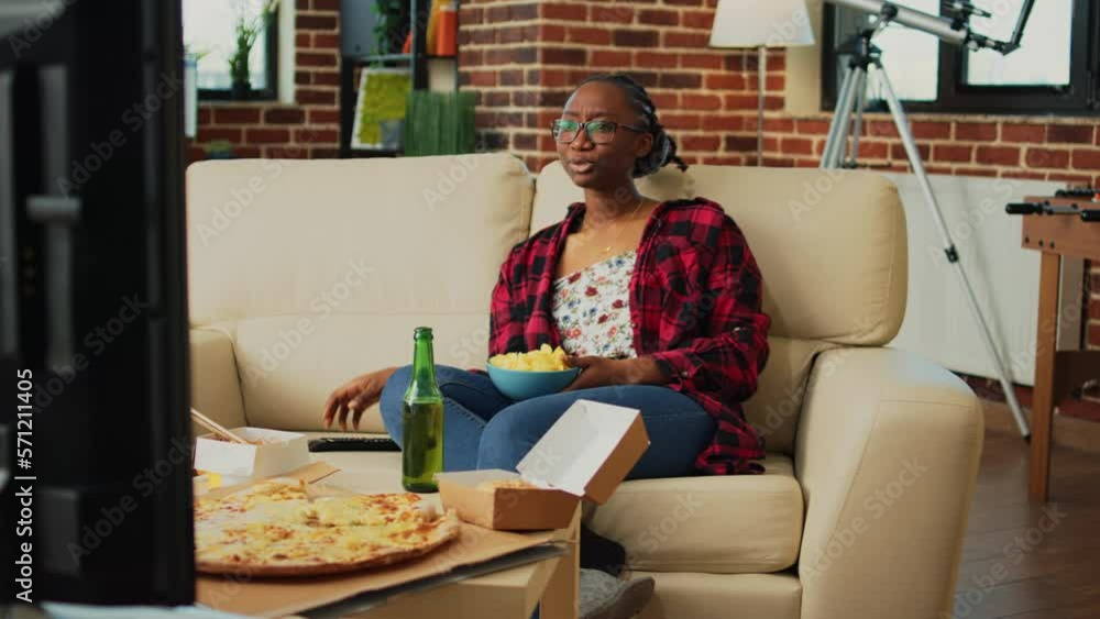 Happy woman laughing at comedy show on television, eating chips and snacks from bowl in living room. Smiling modern girl serving multiple fast food meals from takeout, drinking beer. Happy woman laughing at comedy show on television, eating chips and snacks from bowl in living room. Smiling modern girl serving multiple fast food meals from takeout, drinking beer.