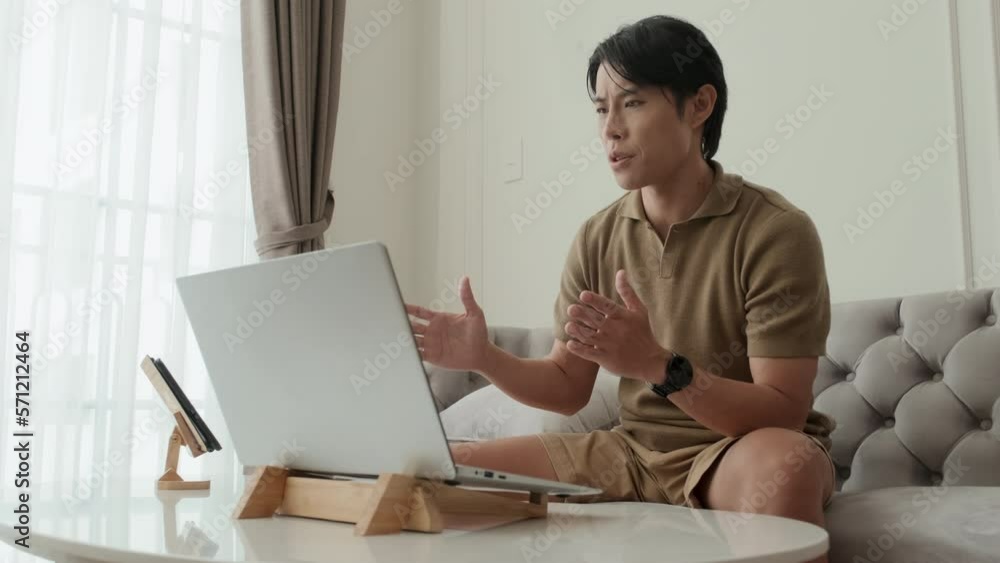 Young confident businessman explaining something to colleague on computer screen during digital meeting while sitting on couch in front of laptop at home Young confident businessman explaining something to colleague on computer screen during digital meeting while sitting on couch in front of laptop at home
