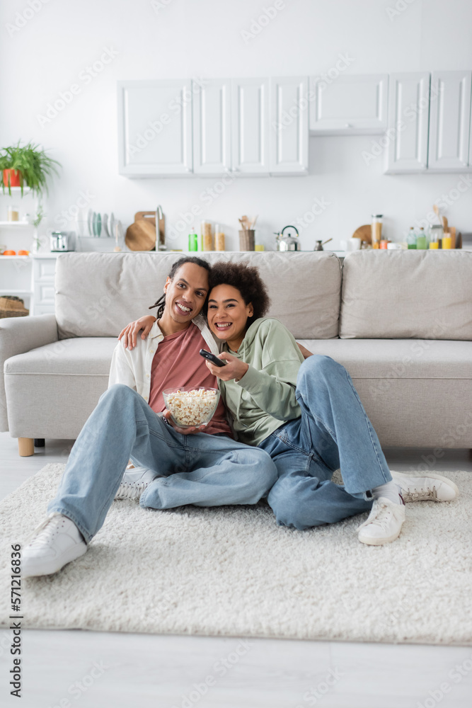 Smiling african american woman hugging boyfriend with popcorn while watching tv at home. Smiling african american woman hugging boyfriend with popcorn while watching tv at home.