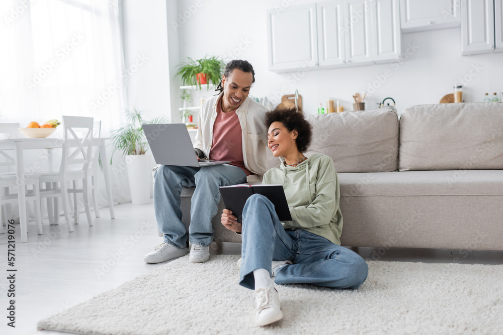 Positive african american woman holding notebook near boyfriend with laptop at home.