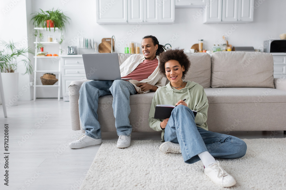 Smiling african american woman writing on notebook near boyfriend with laptop at home. Smiling african american woman writing on notebook near boyfriend with laptop at home.