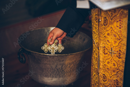 A priest is holding a golden cross necklace and purifying it in water for an orthodox baby child baptism.