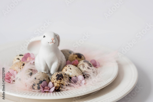 Banner. The concept of a bright holiday is a white rabbit and Easter quail eggs with pink flowers and feathers in a plate on a white isolated background. Beautiful Easter card.