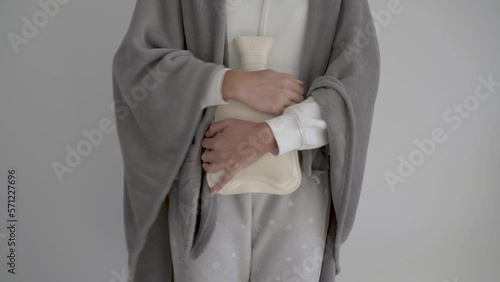 Girl in warm light-colored home clothes and a plaid stands against a white wall, hugging a beige rubber heating pad from the cold. The girl's face is out of frame. Medium shot. Cold winter in Europe. 