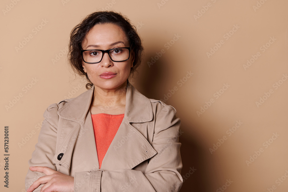Confident middle-aged pretty woman, wearing stylish eyeglasses, poses with folded arms over beige background. Copy space