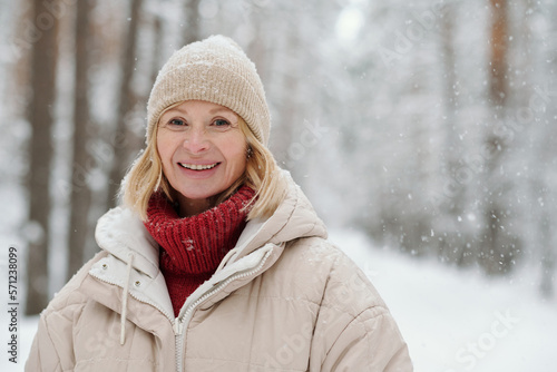 Happy mature blond woman in woolen knitted beanie, red sweater and warm winter jacket looking at camera in forest during snowfall