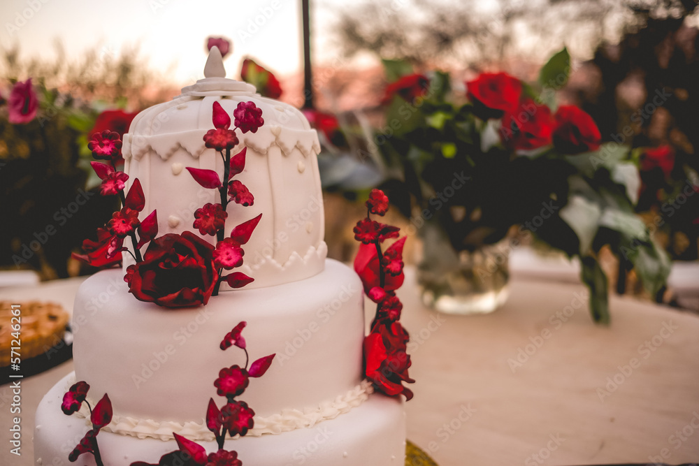 Beautiful white wedding cake with red roses and flowers made of sugar decoration in a wedding table at sunset Beautiful white wedding cake with red roses and flowers made of sugar decoration in a wedding table at sunset