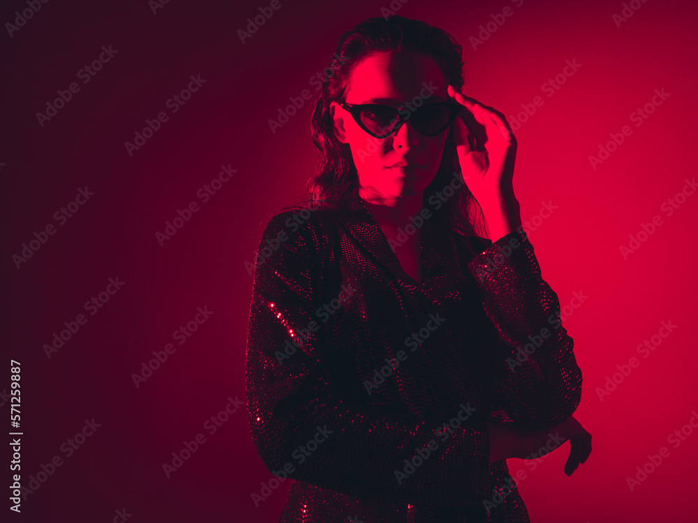 Young beautiful confident brunette female in trendy evening shiny green reflecting dress. Sexy carefree woman isolated on red neon light in studio. Fashionable model in sunglasses. high fashion Young beautiful confident brunette female in trendy evening shiny green reflecting dress. Sexy carefree woman isolated on red neon light in studio. Fashionable model in sunglasses. high fashion