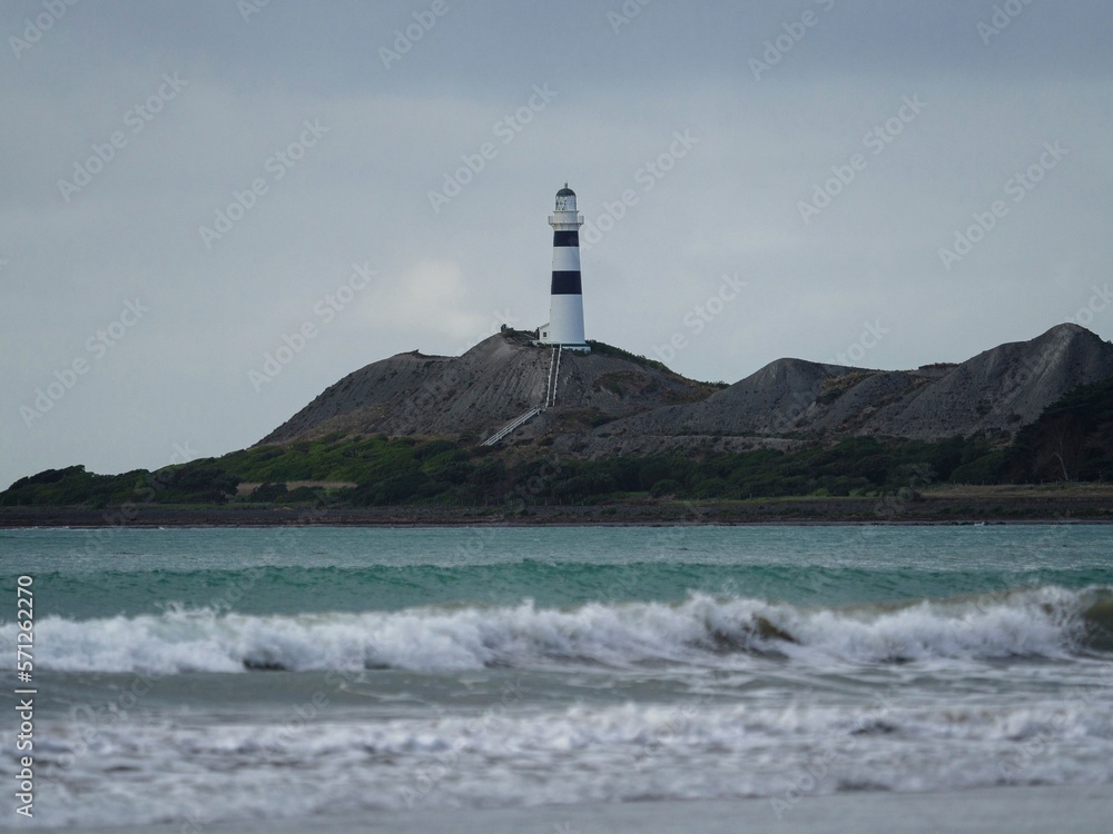 Waves crashing onto ocean beach with black and white Cape Campbell lighthouse in background, Marlborough New Zealand