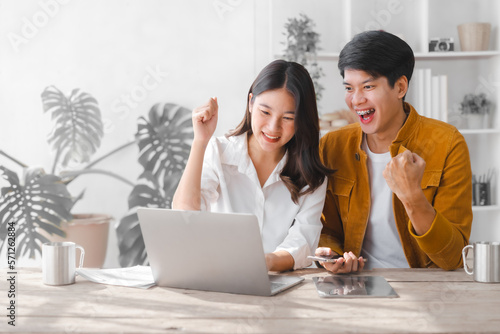 Cheers, Happy Asian couple husband and wife using laptop and paper bills at home use banking applications sit together at the table A cautious family spouse is confused by high expenses.