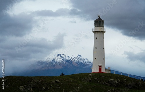 Low angle grass shot of tall white Cape Egmont Lighthouse with Mount Taranki in background North Island New Zealand
