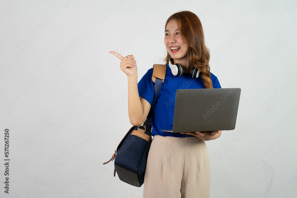 Attractive young Asian college student with a backpack on her shoulder making a gesture with pointing hand and holding a laptop in her hand. isolated on white background Attractive young Asian college student with a backpack on her shoulder making a gesture with pointing hand and holding a laptop in her hand. isolated on white background