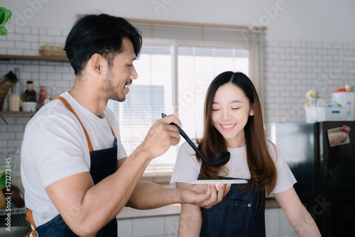 Asian young man feeding pumpkin soup to girlfriend in kitchen.