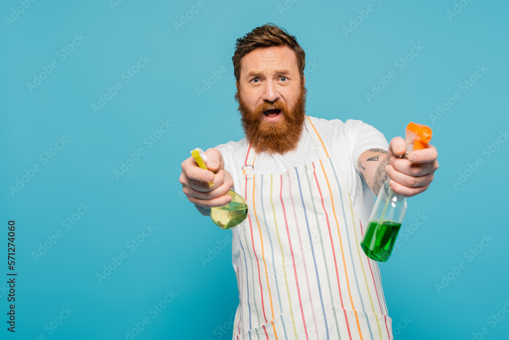 excited man in apron having fun while spraying detergents at camera isolated on blue. excited man in apron having fun while spraying detergents at camera isolated on blue.