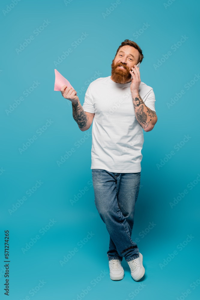 full length of happy bearded man in white t-shirt and jeans holding rag and talking on cellphone on blue background. full length of happy bearded man in white t-shirt and jeans holding rag and talking on cellphone on blue background.