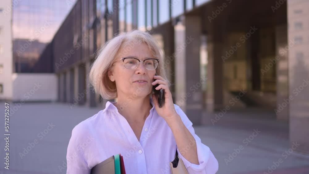 Smiling professional mature mature old business woman wears glasses using using talking phone smartphone street outdoor. Happy senior grey-haired older employee 50s 60s businesswoman working on phone Smiling professional mature mature old business woman wears glasses using using talking phone smartphone street outdoor. Happy senior grey-haired older employee 50s 60s businesswoman working on phone