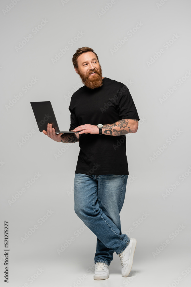 full length of positive bearded man in jeans standing with laptop and looking away on grey background.