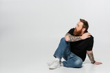 full length of tattooed man in blue jeans and white sneakers sitting and looking away on grey background.