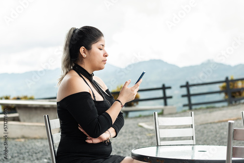 young latina woman, looking at her cell phone, while sitting in an outdoor restaurant, waiting