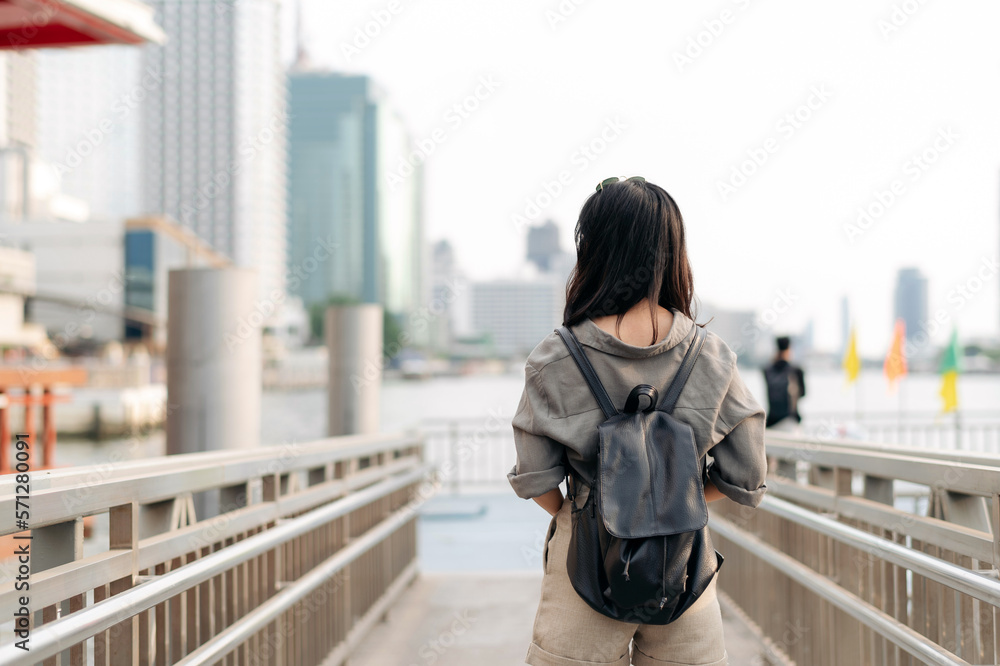 Young Asian woman backpack traveler waiting an express boat at pier on Chao Phraya River in Bangkok. Journey trip lifestyle, world travel explorer or Asia summer tourism concept Young Asian woman backpack traveler waiting an express boat at pier on Chao Phraya River in Bangkok. Journey trip lifestyle, world travel explorer or Asia summer tourism concept