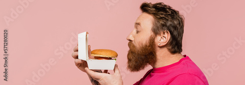 Tela side view of bearded man smelling delicious hamburger isolated on pink, banner