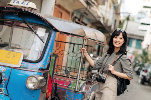 Young Asian woman backpack traveler standing a side of Tuk Tuk taxi on summer vacations at Bangkok, Thailand. 