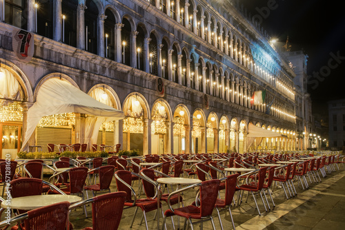 Venice, Italy empty Saint Mark square with arcades at night. Illuminated view of closed stores with empty red chairs stacked around tables, without crowd at iconic Piazza San Marco.