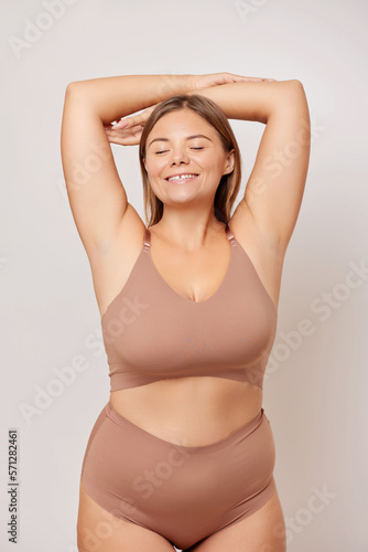 Body positivity and self love concept. Waist up shot of joyful body positive young woman keeps arms raised feels confident love own body wears beige lingerie poses over white wall