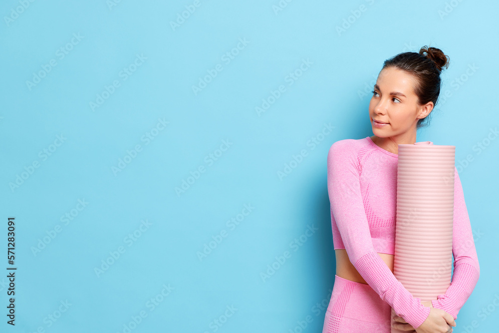 Smiling slim woman in pink tracksuit looks aside to the copy space poses with rolled mat does yoga pilates or fitness isolated over blue background. Advertisement concepts Smiling slim woman in pink tracksuit looks aside to the copy space poses with rolled mat does yoga pilates or fitness isolated over blue background. Advertisement concepts