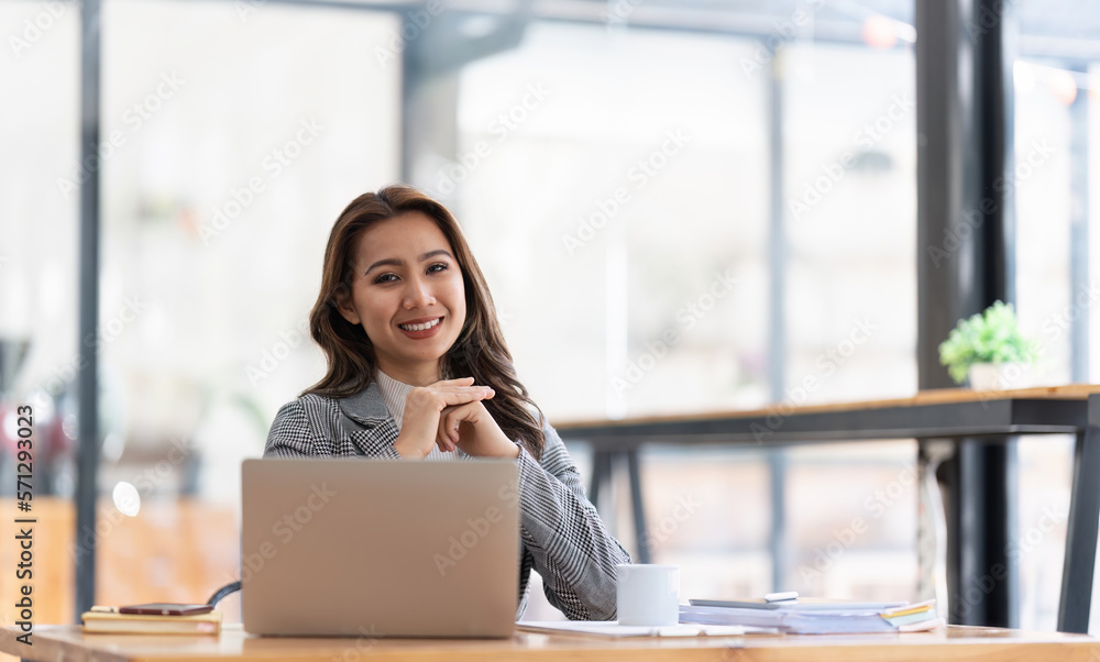 Young businesswoman working at her laptop and going over paperwork while sitting at a desk in an office Young businesswoman working at her laptop and going over paperwork while sitting at a desk in an office
