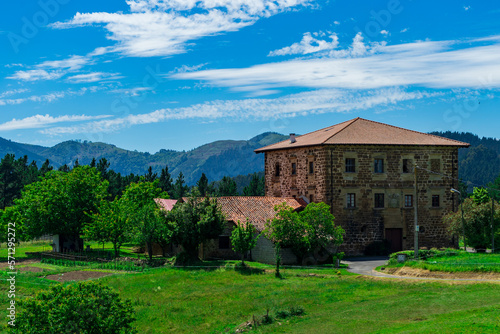 An old brick manor with 3 floors and a tiled roof against the backdrop of mountains and green grass. Basque Country, Spain