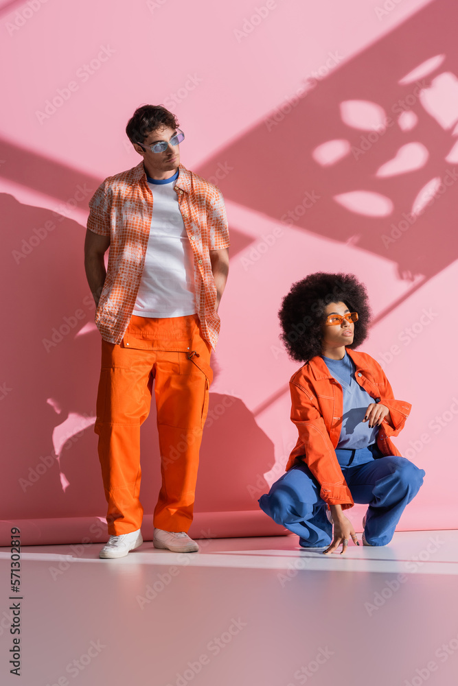 Obraz premium Interracial couple in sunglasses looking away on pink background with shadow.